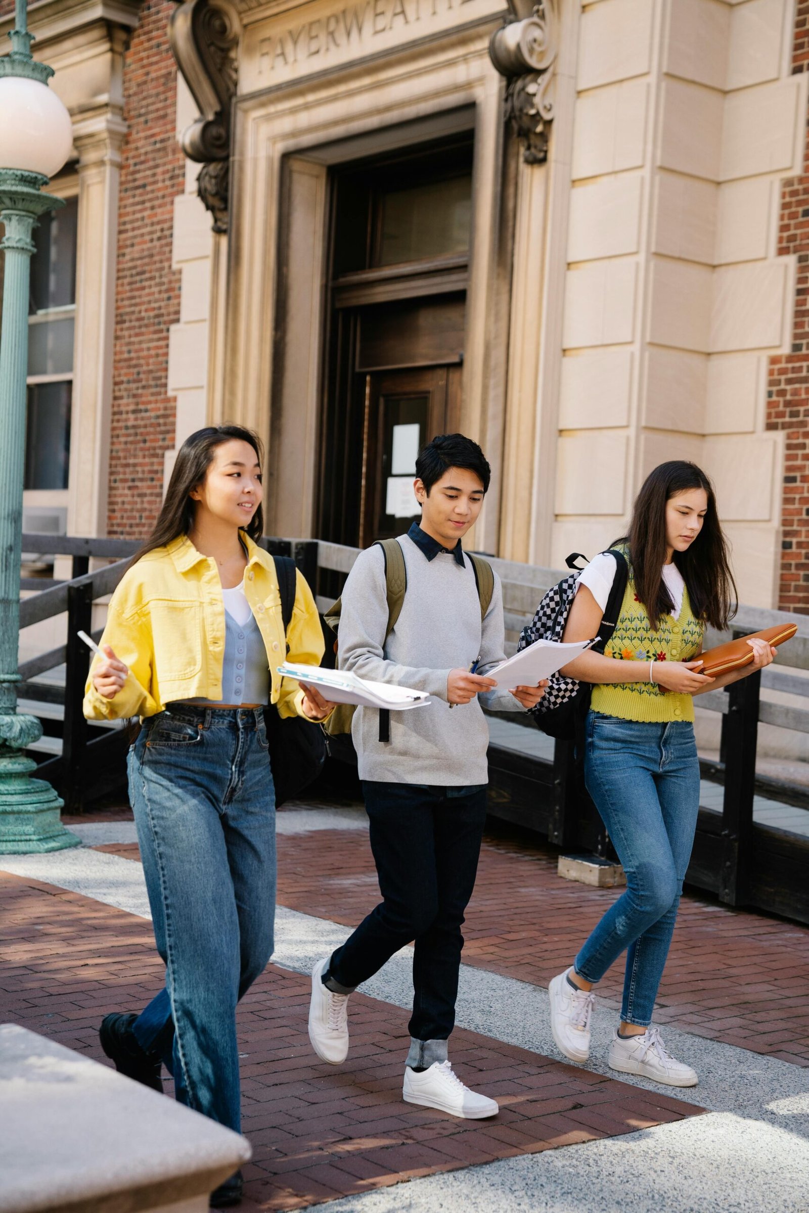 Three diverse students walking together outdoors on a university campus, carrying books and papers.